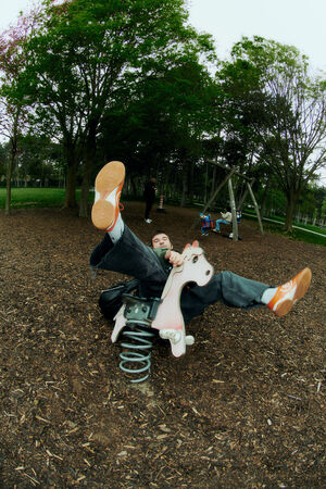 A person in casual clothes, carrying an INA KENT bag, is falling backward off a spring-mounted playground ride shaped like a horse, with legs in the air and wood chips on the ground.