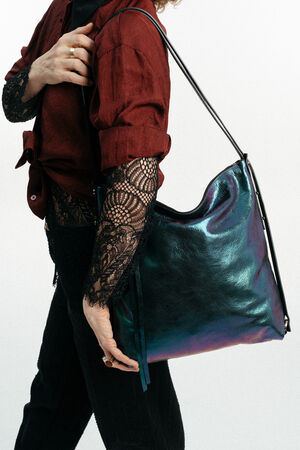 Eine Person, die ein burgunderrotes Hemd und schwarze Spitzenärmel trägt, hält eine große, schillernde INA KENT-Umhängetasche vor einem schlichten weißen Hintergrund.