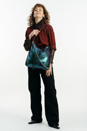 Eine Person mit lockigem Haar steht mit geschlossenen Augen, trägt ein rotes Oberteil, eine schwarze Hose und schwarze Schuhe und hält eine glänzende, schillernde Handtasche von INA KENT mit einem Kettenriemen.