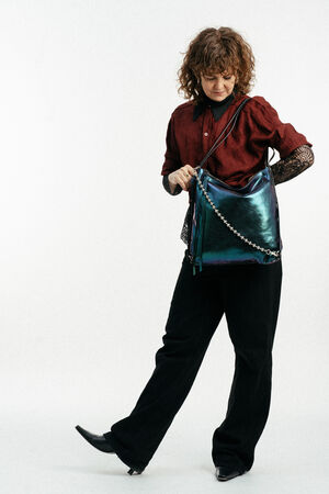 Eine Person mit lockigem Haar, die ein kastanienbraunes Oberteil und eine schwarze Hose trägt, schaut in eine große, glänzende, dunkel schillernde INA KENT-Handtasche mit einem Kettenriemen.