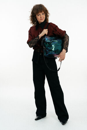 Eine Person mit lockigem Haar, die ein kastanienbraunes Hemd mit Spitzenärmeln und eine schwarze Hose trägt, hält eine glänzende, schillernde INA KENT-Tasche vor einem schlichten weißen Hintergrund.