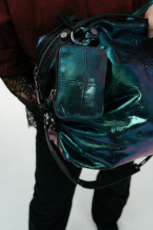 Person mit einer irisierenden blau-grünen INA KENT-Handtasche, an der ein passender kleiner Beutel befestigt ist, bekleidet mit einem roten Oberteil und einer schwarzen Hose.