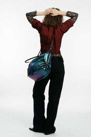 Person, die mit dem Rücken zur Kamera steht, ein rotes Spitzenoberteil und eine schwarze Hose trägt und eine große, schillernde INA KENT-Umhängetasche in der Hand hält, die Arme erhoben und die Hände auf dem Kopf.