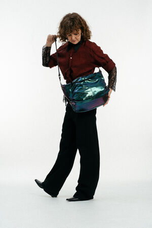 Eine Person mit lockigem Haar trägt ein kastanienbraunes Hemd mit Spitzenärmeln, eine schwarze Hose und schwarze Schuhe und hält eine große schillernde INA KENT-Umhängetasche vor einem weißen Hintergrund.