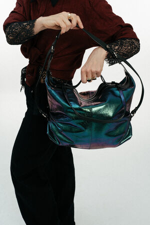 Eine Person, die eine burgunderfarbene Bluse mit Spitzenärmeln trägt, hält mit beiden Händen eine große schillernde INA KENT-Handtasche vor einem schlichten weißen Hintergrund.