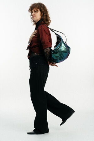 Eine Person mit lockigem Haar, roter Bluse, schwarzer Hose und schwarzen Schuhen trägt eine metallisch blaue Handtasche von INA KENT auf der Schulter und steht vor einem schlichten weißen Hintergrund.