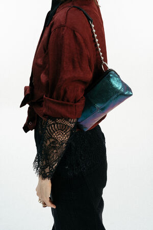 Person, die ein dunkelrotes Hemd mit Spitzenärmeln und eine schwarze Hose trägt und eine kleine schillernde INA KENT-Handtasche mit einem Kettenriemen über der Schulter hält.