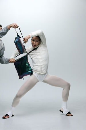 Eine Person in weißer Tanzkleidung hält eine glänzende INA KENT-Tasche, während eine andere Hand versucht, die Tasche wegzuziehen, vor einem schlichten Hintergrund.