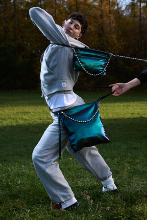 A person in a grey tracksuit poses outdoors while being tugged backward by two metallic teal INA KENT handbags with chain straps, held by someone out of frame.