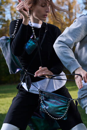 Person outdoors wearing layered clothing and multiple iridescent INA KENT bags with chains, holding black straps while another person in grey moves beside them. Trees are visible in the background.