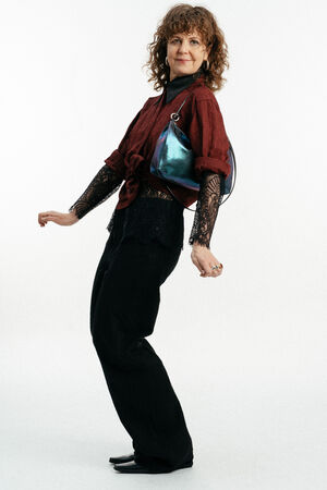 Eine Person mit lockigem Haar steht im Profil, trägt ein rotes Hemd mit schwarzen Spitzenärmeln, eine schwarze Hose und trägt eine INA KENT Metalltasche über der Schulter.