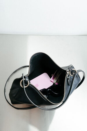 A black INA KENT handbag with a zipper is open on a surface, revealing a pink item, a pair of sunglasses, and some white earphones inside.