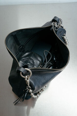 A black INA KENT leather handbag with a chain strap unzipped, revealing a wallet, glasses, and a phone inside, placed on a metallic surface.