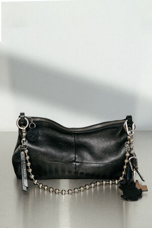 An INA KENT black leather handbag with a silver zipper, metal chain strap, and various keychains and charms attached, placed on a gray surface against a plain background.