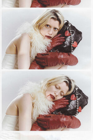 A woman with blonde hair, red lipstick, and red gloves rests her head on an INA KENT black purse decorated with heart-shaped charms while wearing a white feathered accessory.