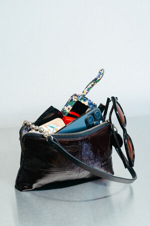 A small, dark INA KENT leather purse sits open on a table, revealing sunglasses, wireless earbuds, lip balm, a patterned keychain, and other personal items inside.