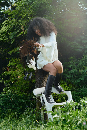 A woman in a white dress and black boots sits on a tree stump atop a broken plastic chair outdoors, reaching towards her brown fringed INA KENT bag.