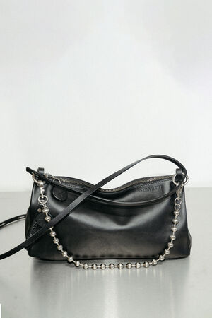An INA KENT black leather handbag with a zipper closure, thin shoulder strap, and a decorative silver beaded chain is displayed on a plain surface.