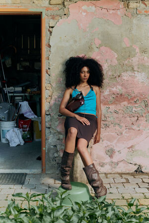 A woman with voluminous dark curly hair wears a blue halter top and brown skirt, leaning against a weathered pink and beige brick wall, her INA KENT bag effortlessly completing the chic look.