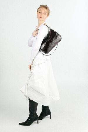 Person wearing a white long-sleeve top, white skirt, black knee-high boots, and carrying a shiny black INA KENT bag over their shoulder, standing against a plain white background.