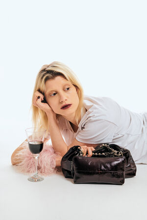 A woman in a white shirt lies on her side next to a glass of red wine, a pink fluffy object, and an INA KENT dark handbag on a white background.