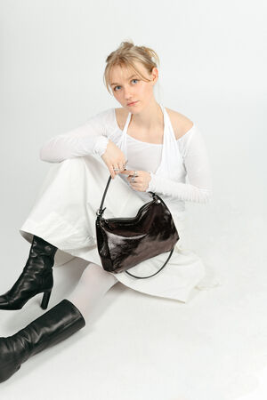 A woman in a white outfit sits on the floor holding an INA KENT black shoulder bag and wearing black heeled boots against a white background.