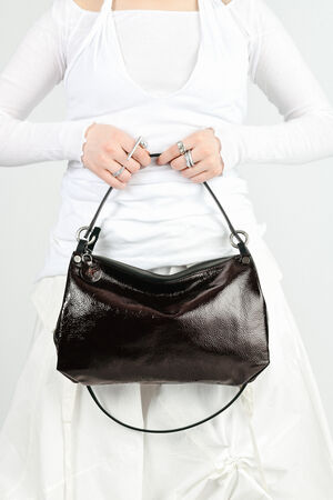 Person in a white outfit holds an INA KENT dark, shiny leather handbag with a zipper and a short strap, displaying it in front of their waist.