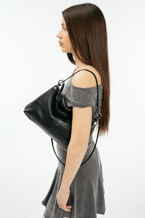 A woman in a grey off-shoulder dress stands in profile, holding an INA KENT black handbag over her shoulder against a plain white background.