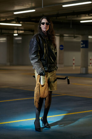 A woman wearing sunglasses, a leather jacket, skirt, and heels stands in a dimly lit parking garage, holding an INA KENT fringed bag.