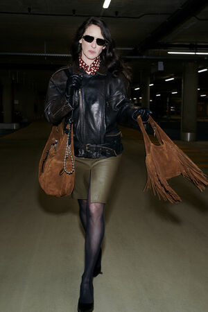 A woman in a leather jacket, olive skirt, black tights, and sunglasses walks through a parking garage holding two brown INA KENT bags, one adorned with fringe.