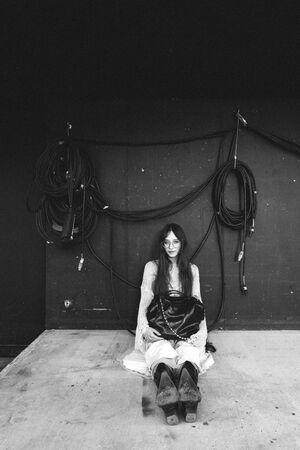 A woman in glasses and boots sits on the ground against a dark wall with coiled cables hanging behind her, an INA KENT bag by her side.