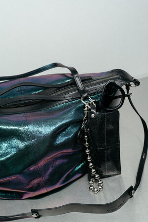Eine schwarze, schillernde INA KENT-Handtasche mit Reißverschluss, schwarzem Trageriemen, einer Sonnenbrille und einem Schlüsselanhänger mit Metallperlen, präsentiert auf einem grauen Untergrund.