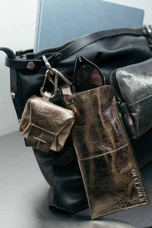 Eine schwarze Ledertasche von INA KENT mit einem blauen Laptop, einer Sonnenbrille in einem metallischen Etui und einem kleinen metallischen Beutel, der am Riemen befestigt ist.