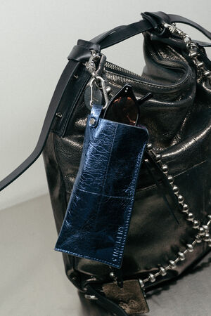 A metallic blue eyeglass case with sunglasses inside is clipped to a black leather INA KENT bag featuring chic chain details.