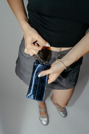 Eine Person, die ein schwarzes Oberteil und einen grauen Rock trägt, hält eine Sonnenbrille und ein glänzendes blaues INA KENT-Etui in der Hand; dabei sind silberne Schuhe zu sehen.