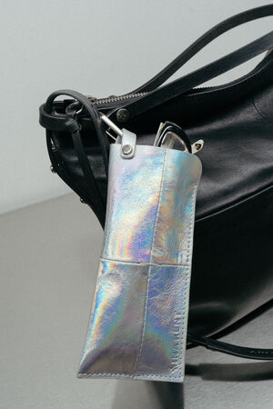 Am Griff einer schwarzen INA KENT-Ledertasche ist mit einem Metallkarabiner ein holografisches Sonnenbrillenetui befestigt, in dem sich eine Sonnenbrille befindet.