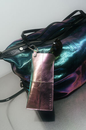 Eine metallisch schillernde INA KENT-Tasche mit schwarzen Trägern und Reißverschluss, an deren Henkel ein glänzendes rosa Brillenetui und eine Sonnenbrille befestigt sind.