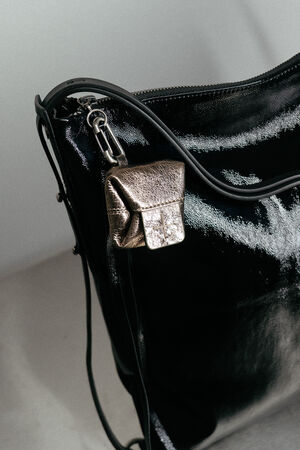 Eine Nahaufnahme einer glänzenden schwarzen INA KENT-Tasche, an deren Seite ein kleines Metalltäschchen mit einem Metallverschluss befestigt ist.