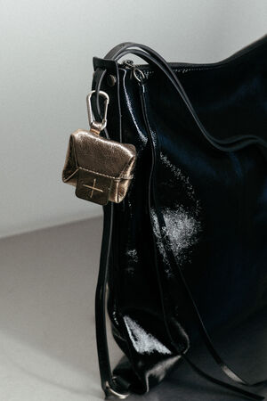 Eine glänzende schwarze INA KENT-Handtasche mit einem goldfarbenen Mini-Täschchen, das mit einem silbernen Verschluss am Henkel befestigt ist, vor einem neutralen Hintergrund.