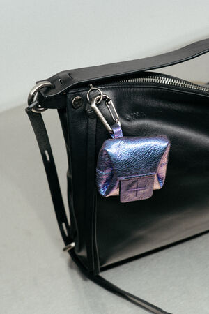 Am Riemen einer schwarzen Ledertasche ist ein kleines, schillerndes, violettes INA KENT-Täschchen mit einem Metallverschluss befestigt.