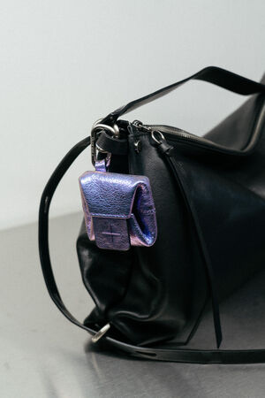 An dem Reißverschluss einer großen schwarzen Ledertasche, die auf einer reflektierenden Oberfläche steht, ist ein metallisch-violetter INA KENT-Beutel befestigt.