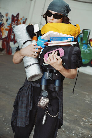 A person wearing sunglasses and a headscarf carries camping gear, books, snacks, and a sleeping bag, with binoculars and INA KENT accessories clipped to their belt, standing in front of graffiti.