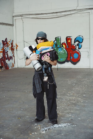 A person wearing dark clothes and sunglasses stands indoors holding INA KENT camping gear, with colorful graffiti art on a white wall in the background.