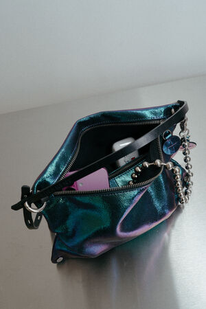 Eine metallisch-blaugrüne INA KENT-Handtasche mit Reißverschluss ist geöffnet und gibt den Blick auf eine Sonnenbrille, ein Handy, einen Schlüsselanhänger und eine Perlenkette frei, die auf einer reflektierenden Oberfläche liegen.