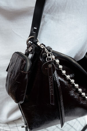 Close-up of a shiny black INA KENT handbag with silver hardware, a small attached pouch, and decorative keychains, worn over a white shirt.