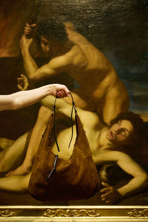 Eine Person hält eine braune Wildlederhandtasche von INA KENT vor einem klassischen Gemälde, das zwei Figuren in einer dramatischen Szene zeigt.