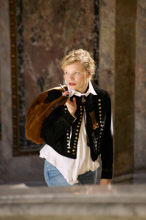 Eine Person mit lockigem blondem Haar trägt eine schwarze, bestickte Jacke, ein weißes Hemd und Jeans; sie hat eine braune INA KENT-Tasche über die Schulter gehängt und steht in einem Innenraum in der Nähe einer Marmorwand.
