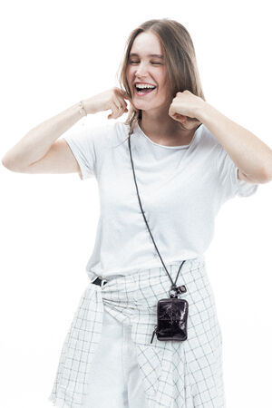 Woman in a white t-shirt and plaid shirt tied at her waist smiles with eyes closed, holding her hands near her face, and wears a small black INA KENT bag on a strap around her neck.