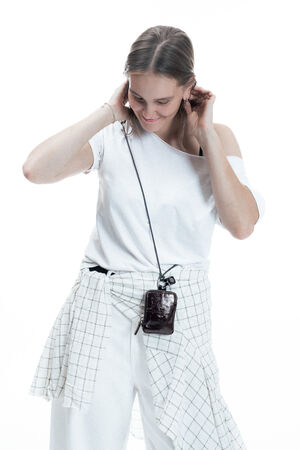 Woman in white outfit with grid-patterned shirt tied at waist, adjusting her hair and wearing a small black INA KENT crossbody pouch, isolated on a white background.