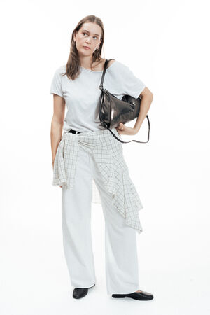 Woman standing against a white background, wearing a white t-shirt, wide-legged pants, checked wrap, black shoes, and holding an elegant INA KENT black shoulder bag.